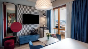 A modern living room with a large flat-screen TV, a sofa, a dining table, and a view of the city through the window. - Hotel Indigo Florence by IHG (Florence)