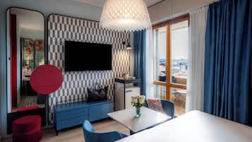 A modern living room with a large flat-screen TV, a sofa, a dining table, and a view of the city through the window.