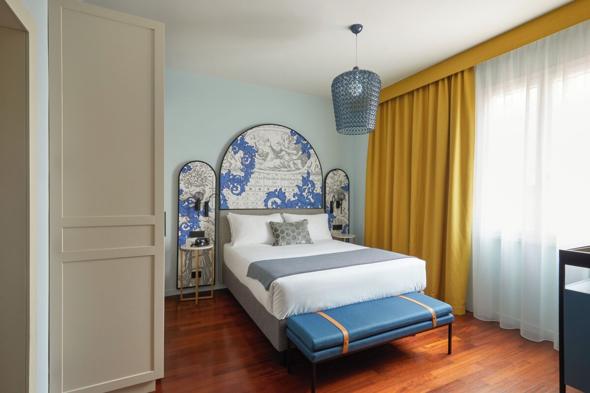 A bedroom with a large bed, a bench, a hanging light fixture, and a view of the outside through sheer curtains.