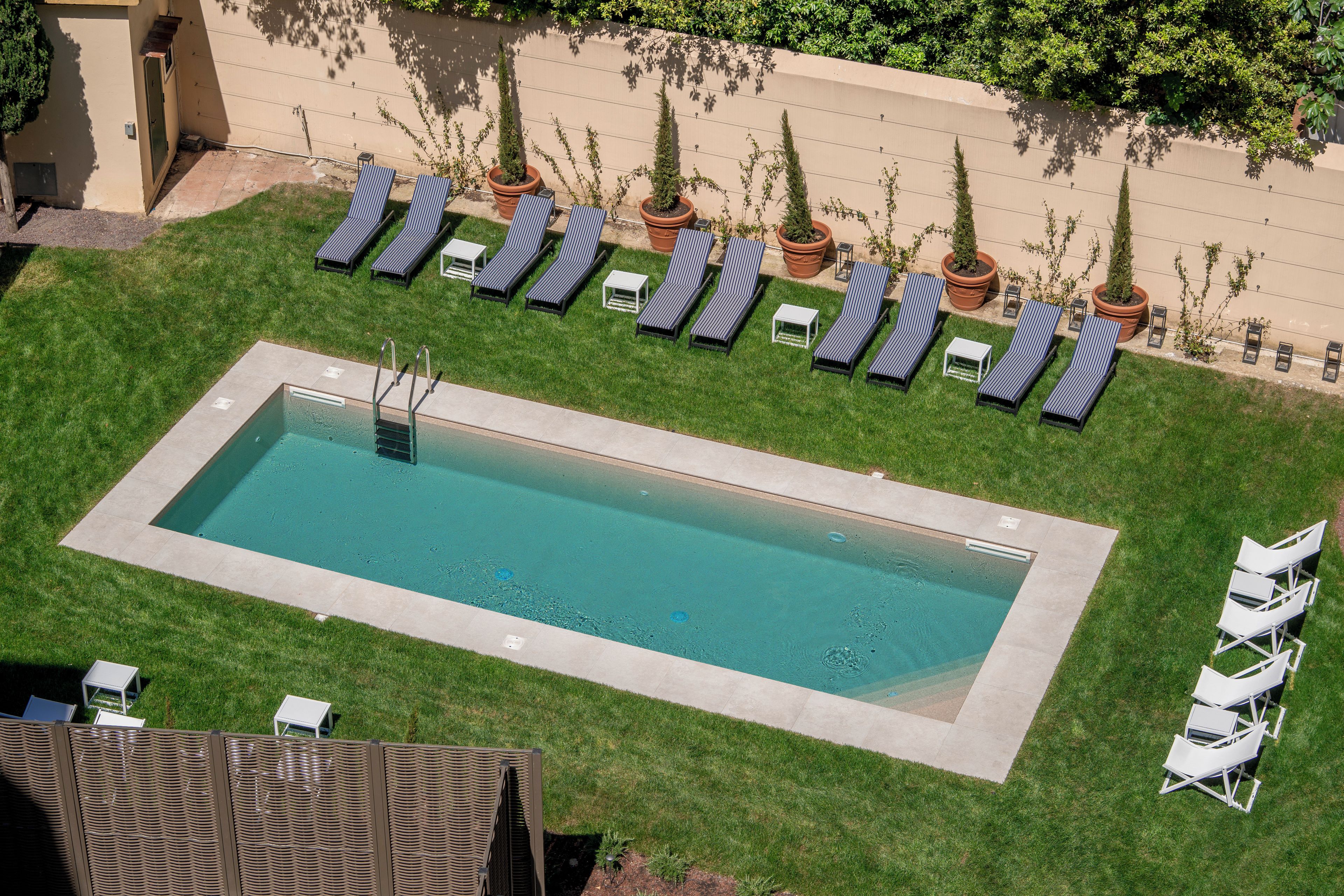 Una piscina rectangular rodeada de sillones de descanso y plantas en macetas, con una vista despejada del cielo.