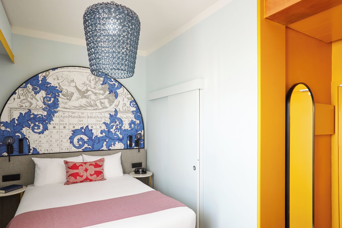 A hotel room with a large bed, a round headboard with a decorative map, a small bedside table with a lamp, and a hanging light fixture.