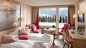 Habitación de hotel con dos camas, una zona de estar con mesa y sillas, y un ventanal con vistas a una montaña nevada.
