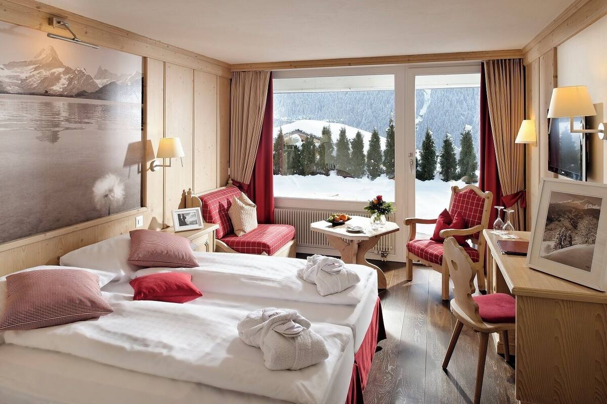 Habitación de hotel con dos camas, una zona de estar con mesa y sillas, y un ventanal con vistas a una montaña nevada.