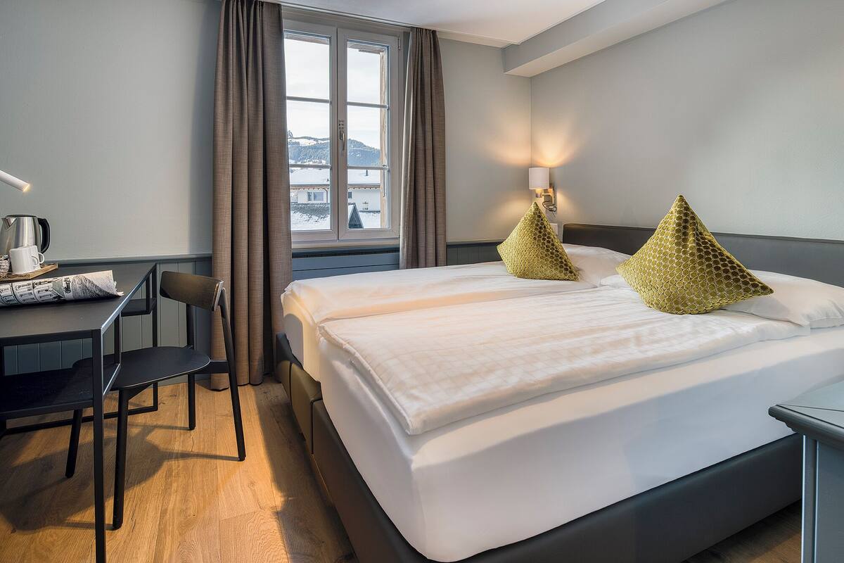 Habitación de hotel con cama, escritorio con silla, ventana con cortinas y lámpara.