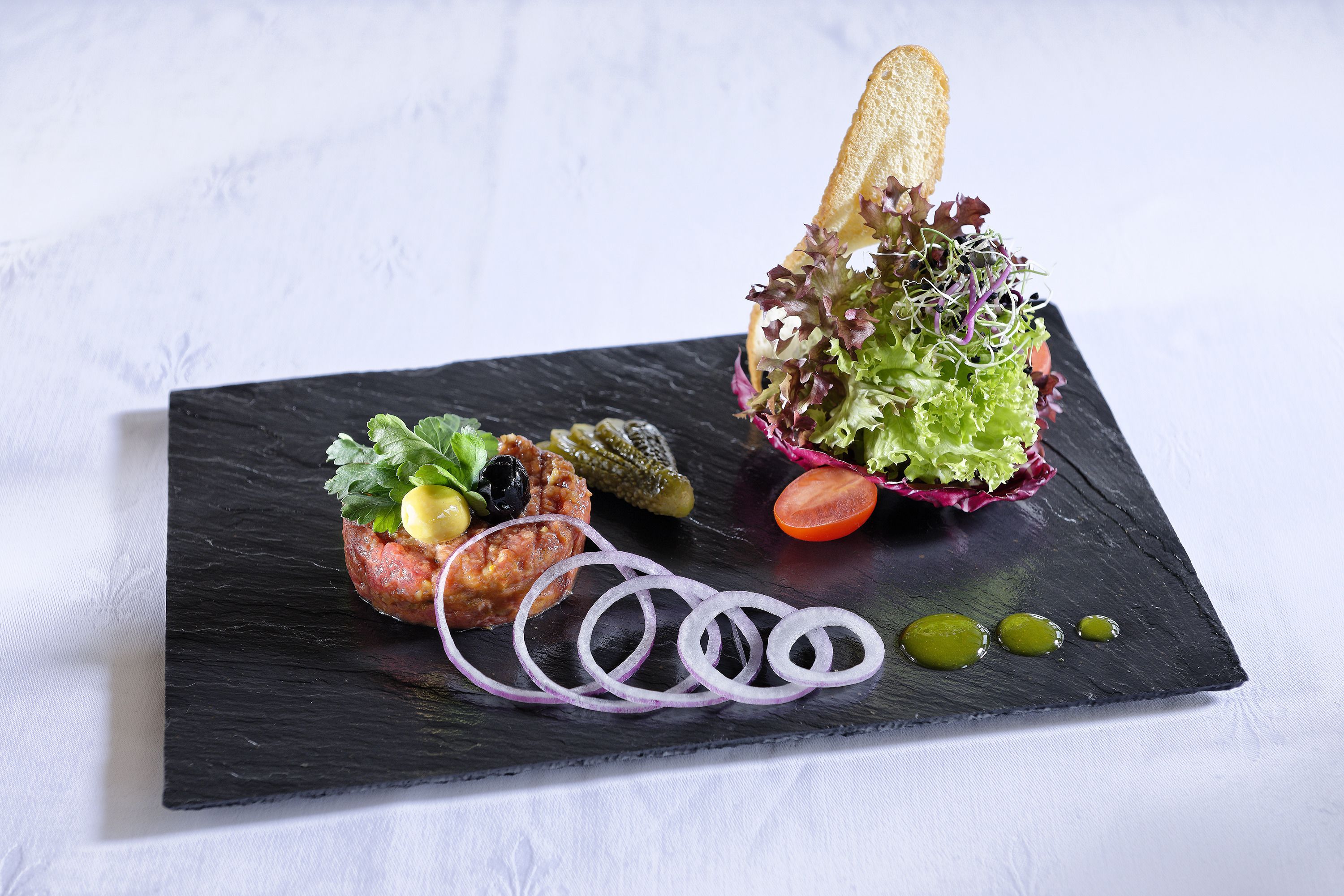 A black slate serving board with a salad, a slice of bread, and a tomato.