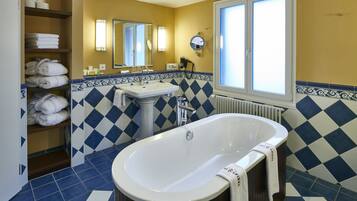 A bathroom with a large bathtub, a sink, and a mirror. There are towels on shelves and a window with blinds.