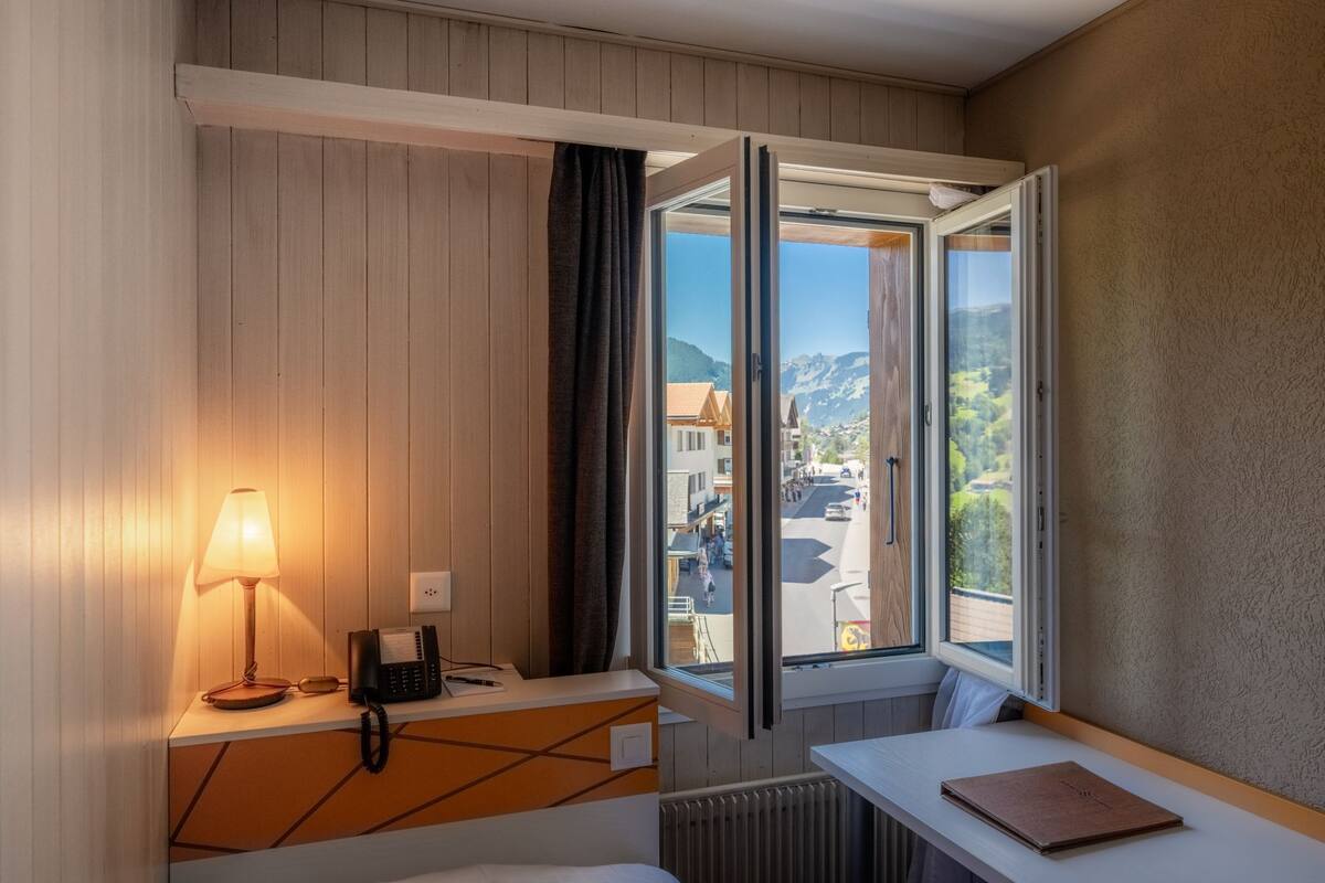 Habitación de hotel con cama, escritorio con lámpara, teléfono y ventana con vista a edificios y montañas.