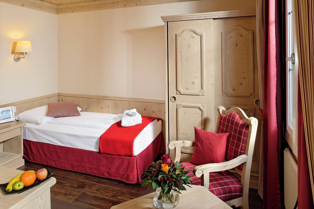 Habitación de hotel con dos camas, un armario de madera, una mesa con frutas y flores, y una silla con cojines rojos.