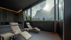 Una habitación moderna con un ventanal grande, dos sillones reclinables, un banco de madera y vistas a las montañas.