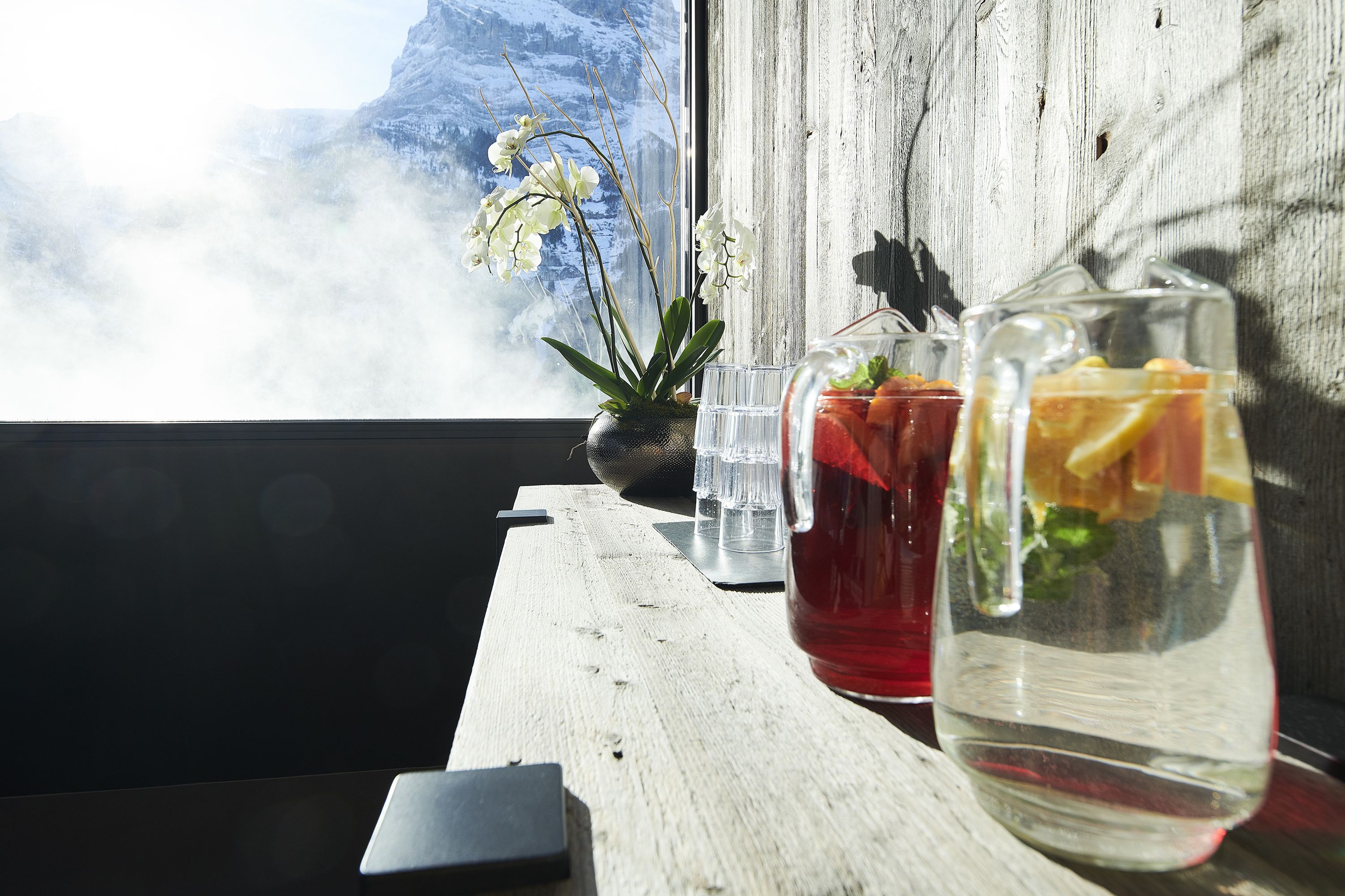 Une table en bois sur laquelle se trouve une carafe en verre remplie d’une boisson et un pot d’orchidée.