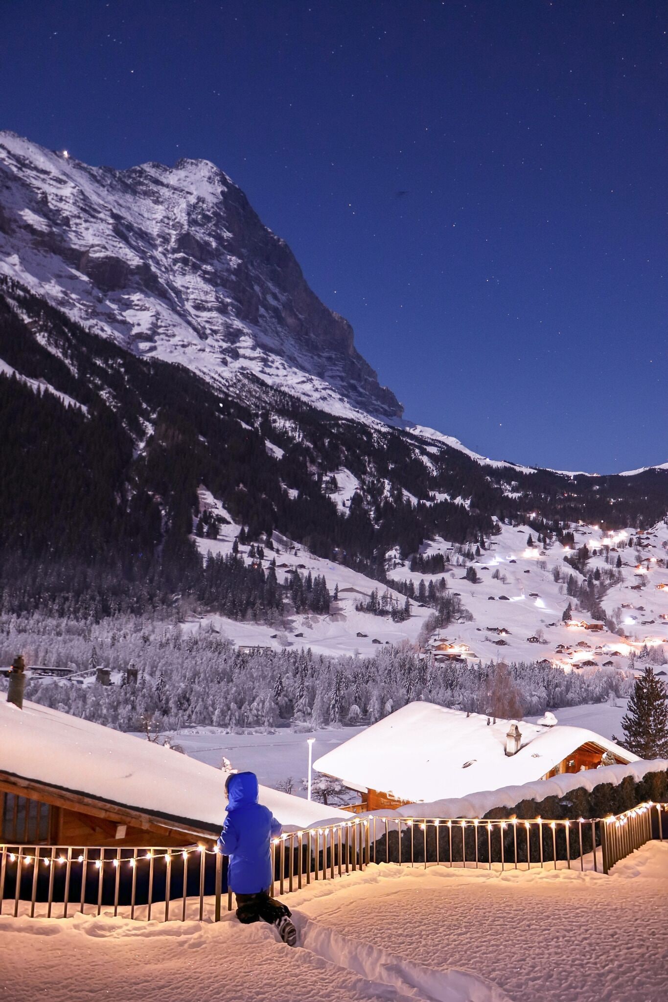 Eine verschneite Berglandschaft bei Nacht, eine Person in einer blauen Jacke geht einen mit Schnee bedeckten Weg entlang.