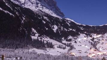 Eine verschneite Berglandschaft bei Nacht, eine Person in einer blauen Jacke geht einen mit Schnee bedeckten Weg entlang.