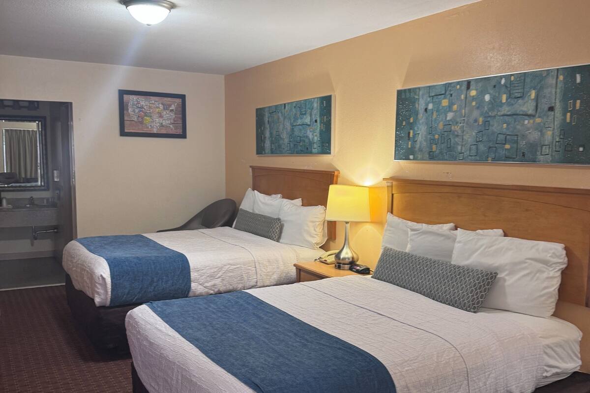 A hotel room with two beds, a nightstand with a lamp, a wooden headboard, and artwork on the walls.