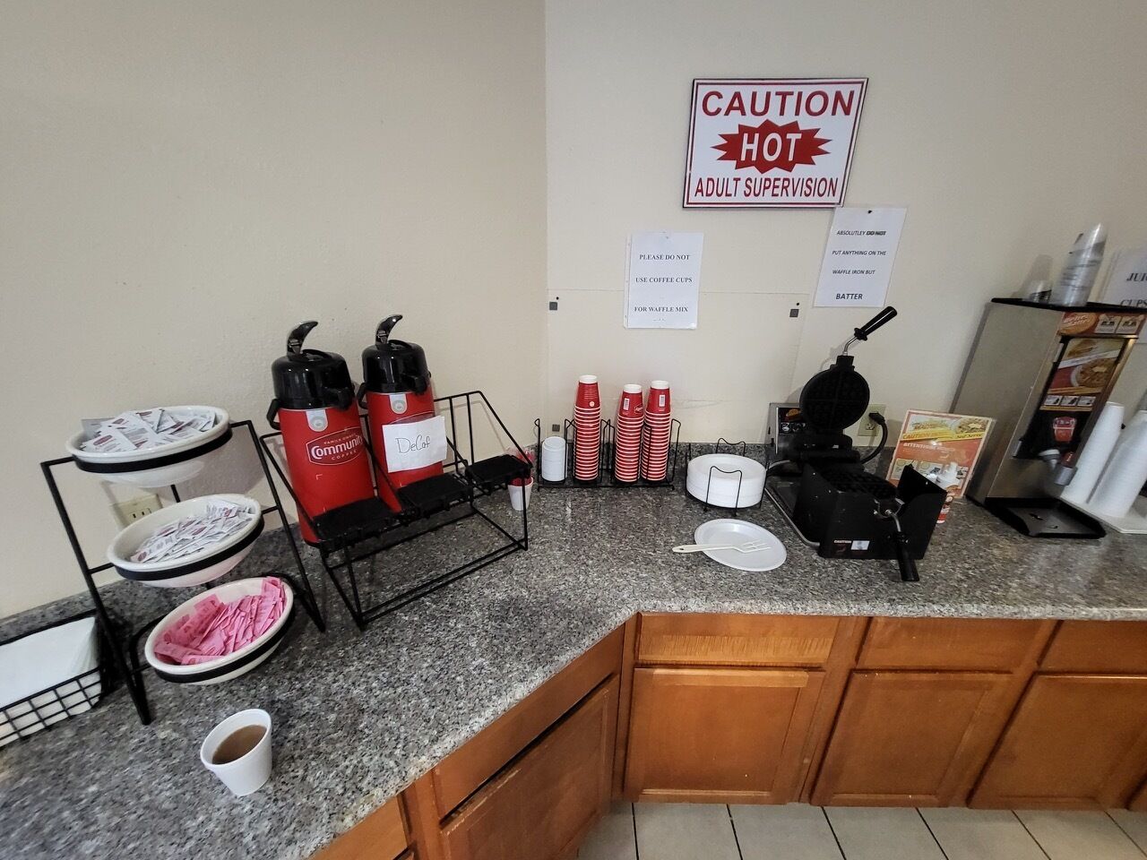 A coffee station with multiple coffee makers, cups, and a waffle iron. There are signs indicating caution and adult supervision.