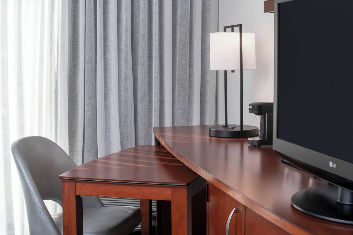 A modern hotel room with a wooden desk, a flat-screen TV, a lamp, and a chair.
