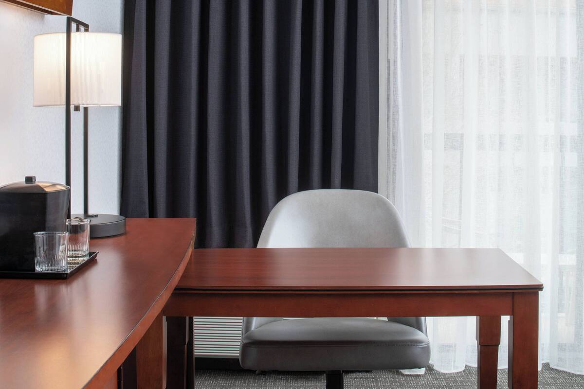A hotel room with a wooden desk, a chair, a lamp, and a window with sheer curtains.