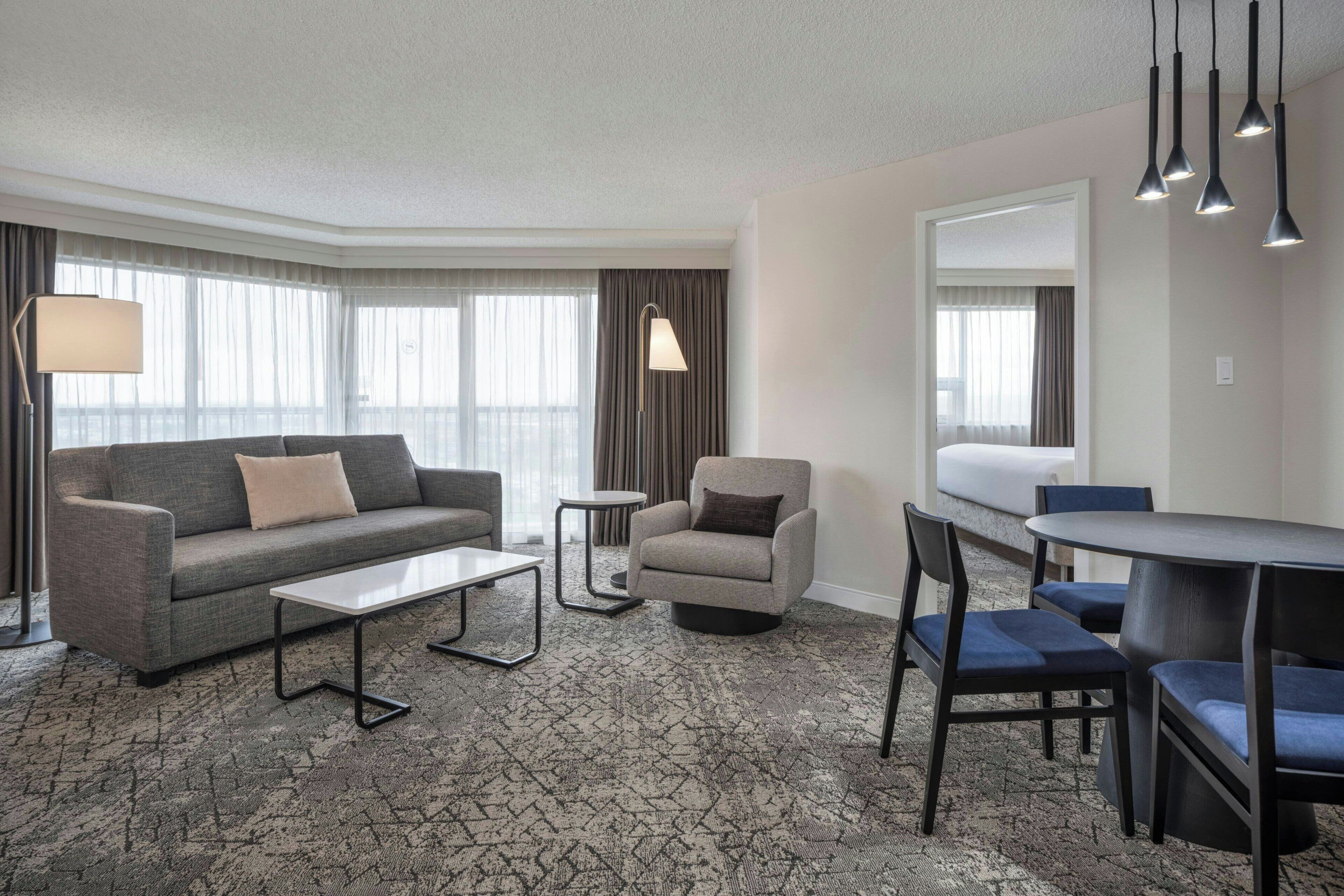 A modern hotel room with a grey sofa, a small round table, a coffee table, a blue chair, and a dining table with chairs.