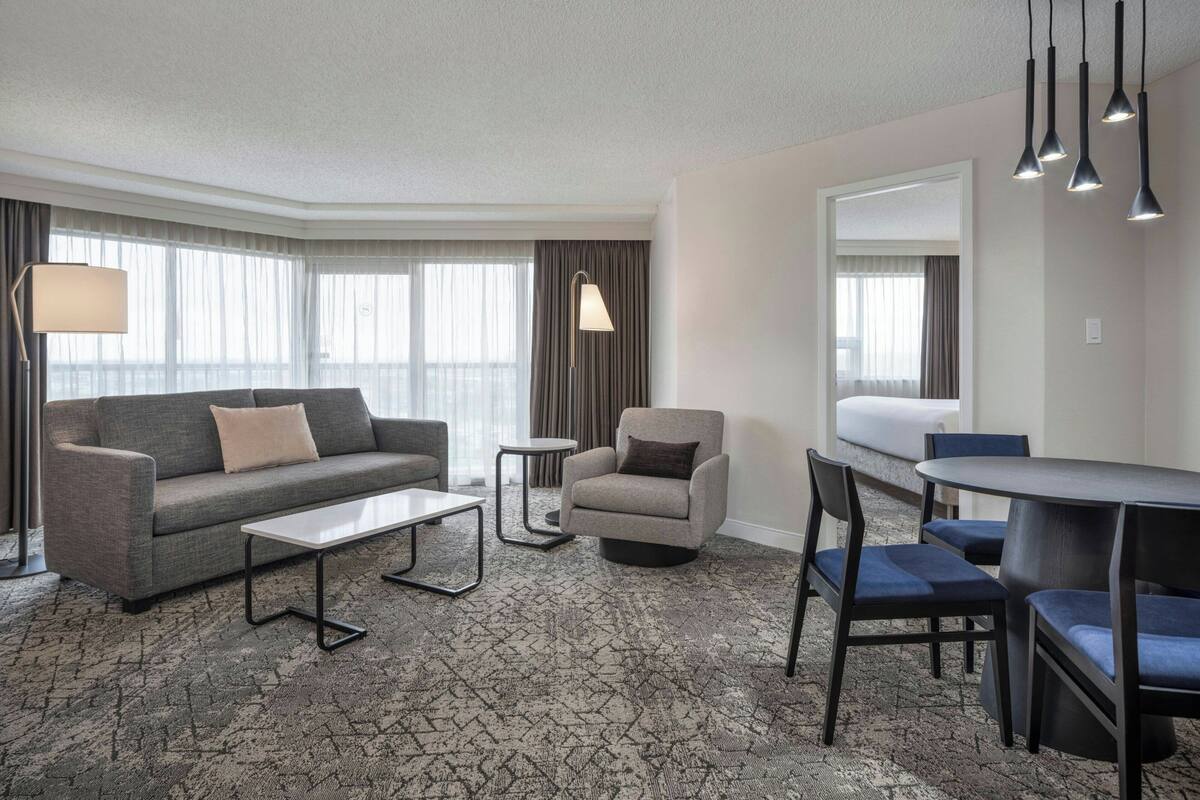 A modern hotel room with a grey sofa, a small round table, a coffee table, a blue chair, and a dining table with chairs.