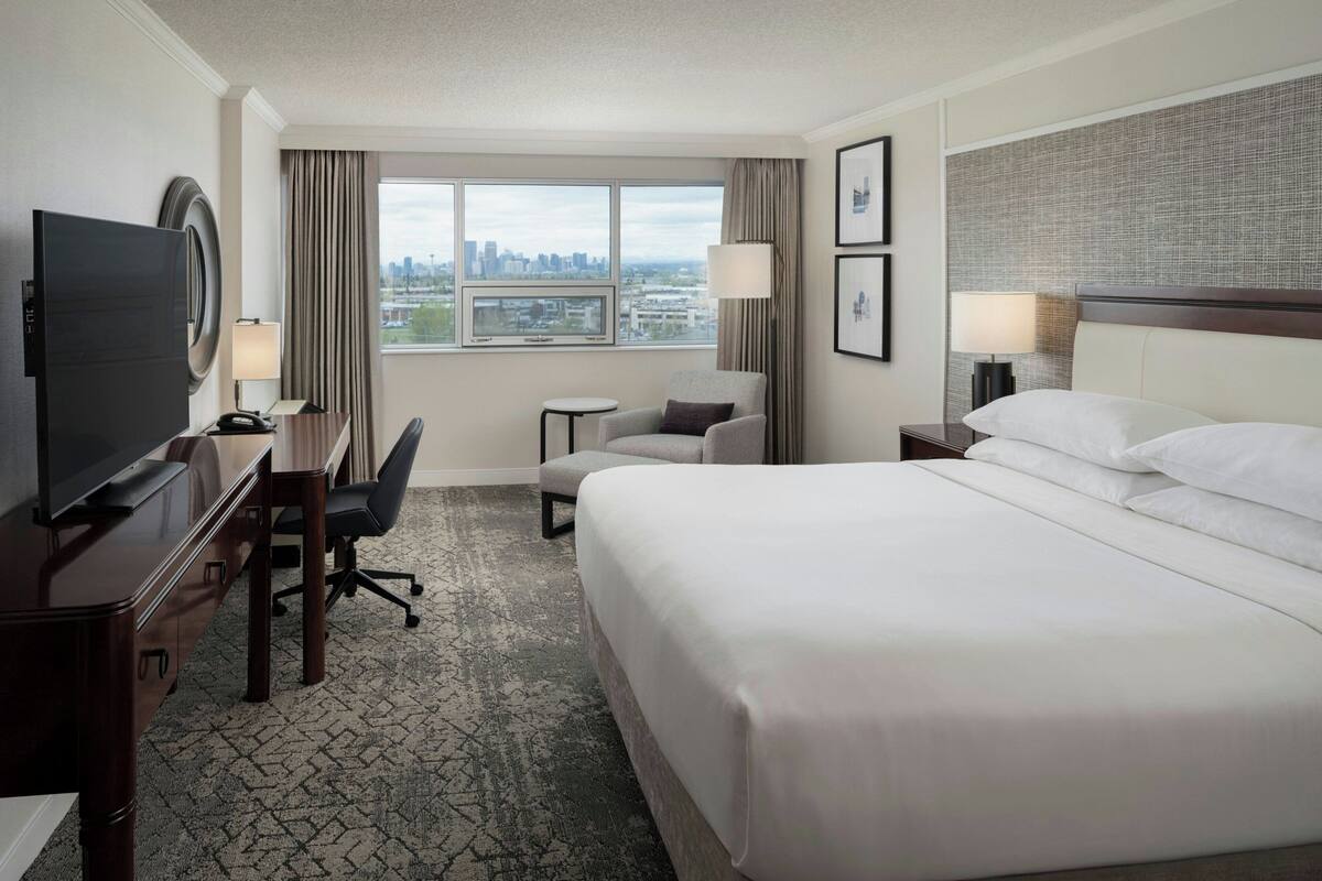 A hotel room with a large bed, a desk with a TV, a chair, a small table, and a view of the city through the window.