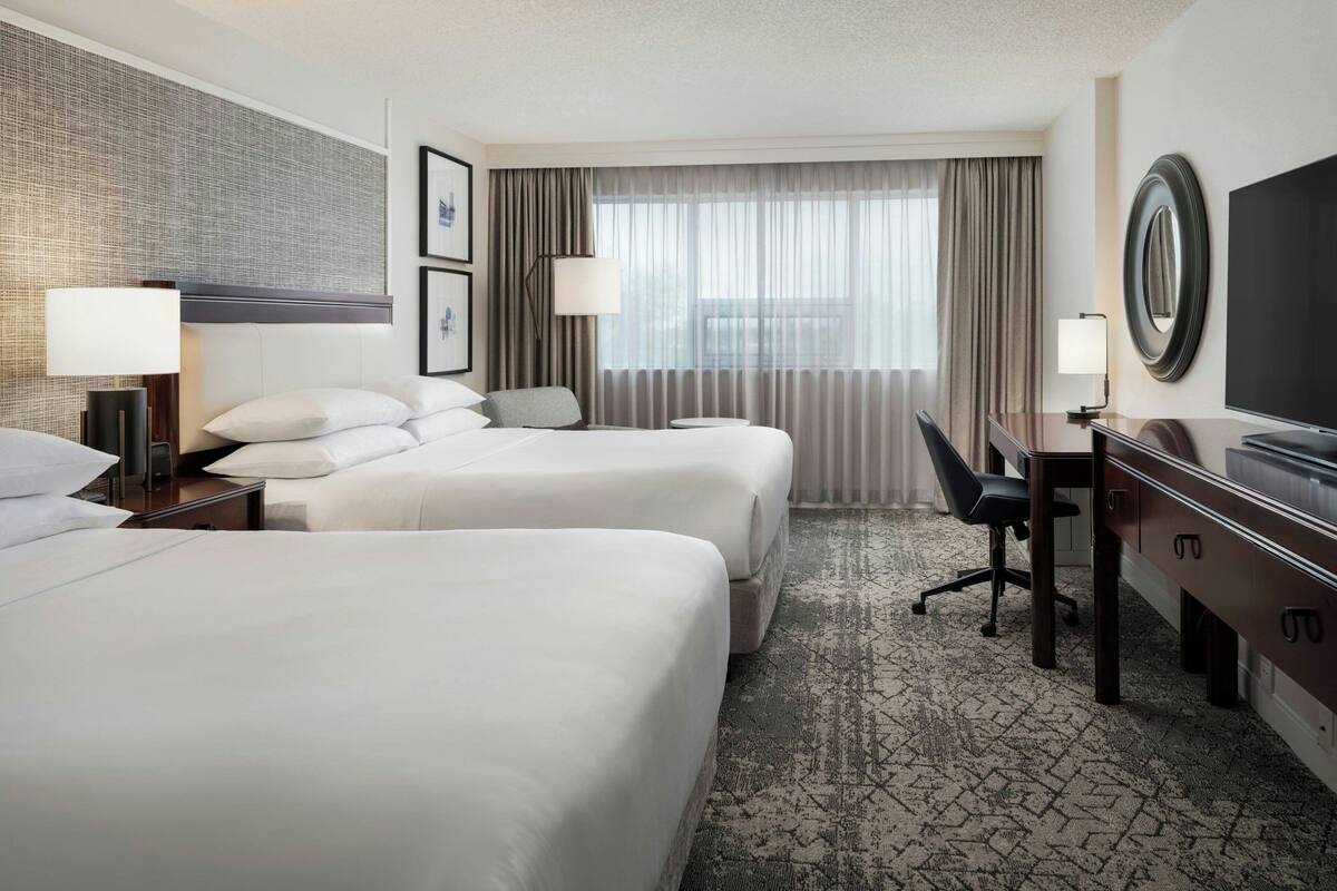 A hotel room with two beds, a desk, a chair, a TV, and a large window with curtains.