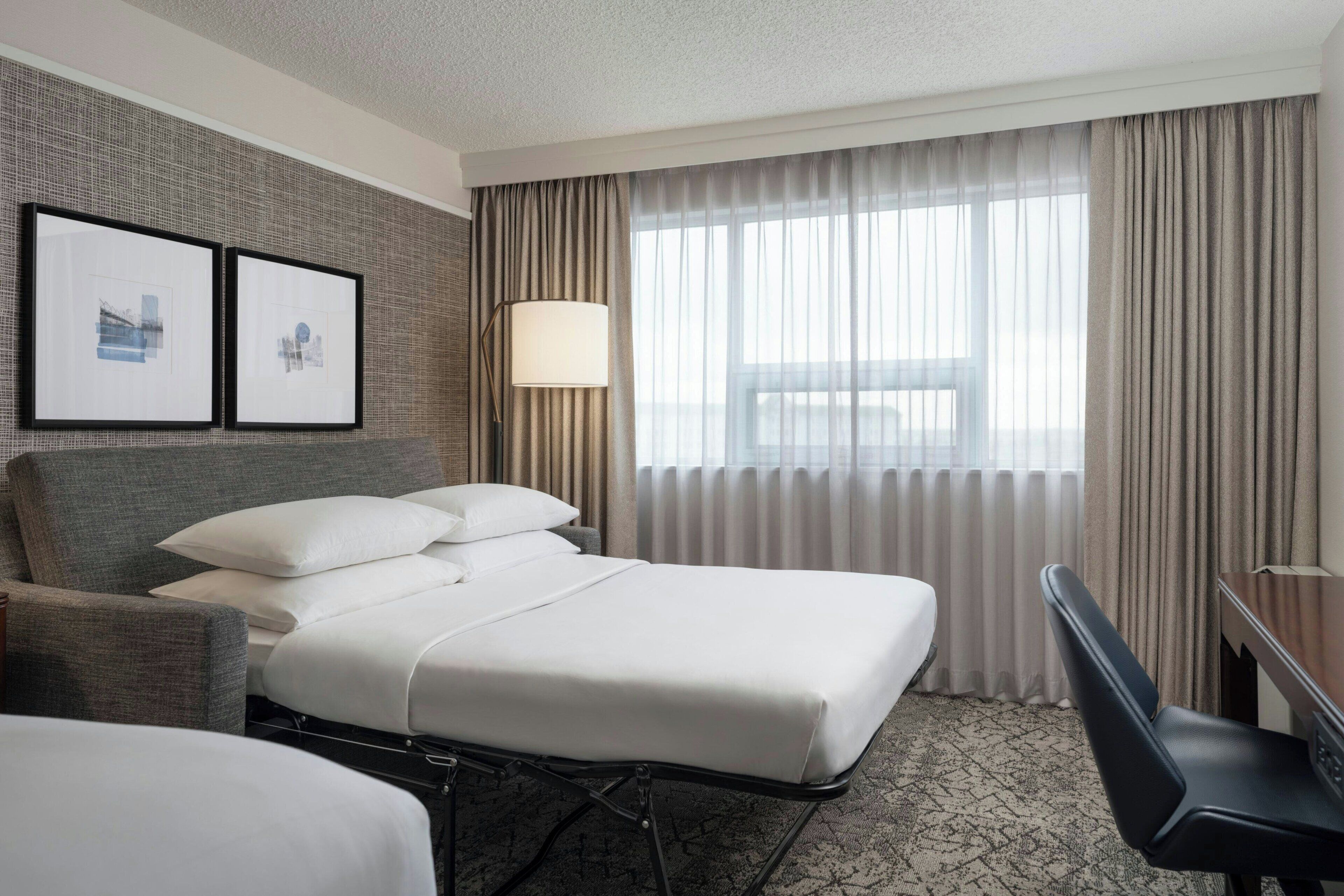A hotel room with a bed, a desk, a chair, and two framed pictures on the wall.