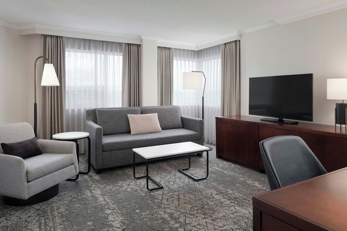 A modern hotel room with a grey sofa, a small table, a TV, and a desk with a chair.