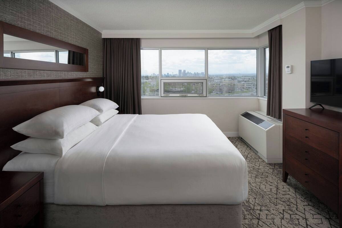 A hotel room with a large bed, a TV, a dresser, and a window with a city view.