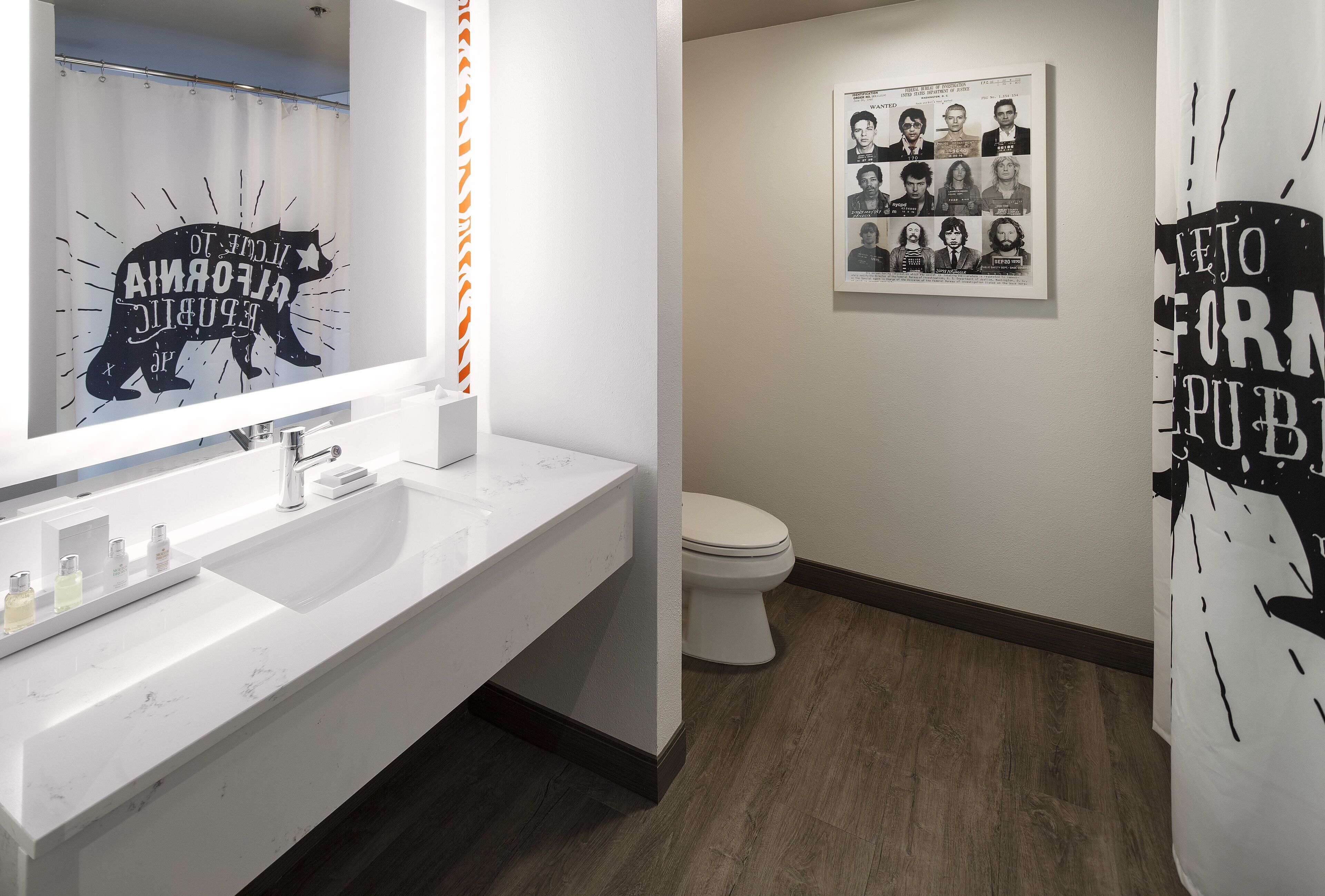 Une salle de bain moderne avec un lavabo blanc, un miroir et des toilettes. Des tableaux encadrés ornent le mur, et il y a un rideau de douche orné d’un motif représentant un ours.