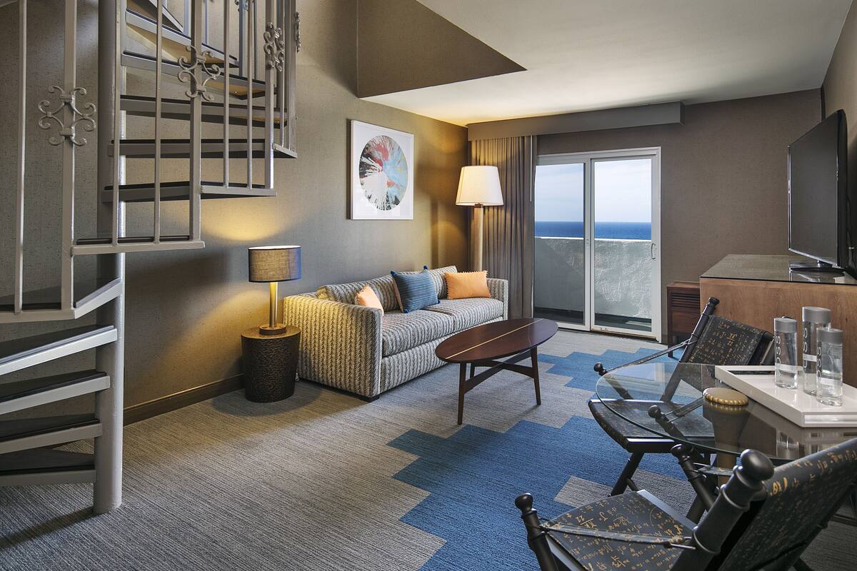A hotel room with a sofa, a table, a chair, a lamp, a TV, and a balcony with a view.