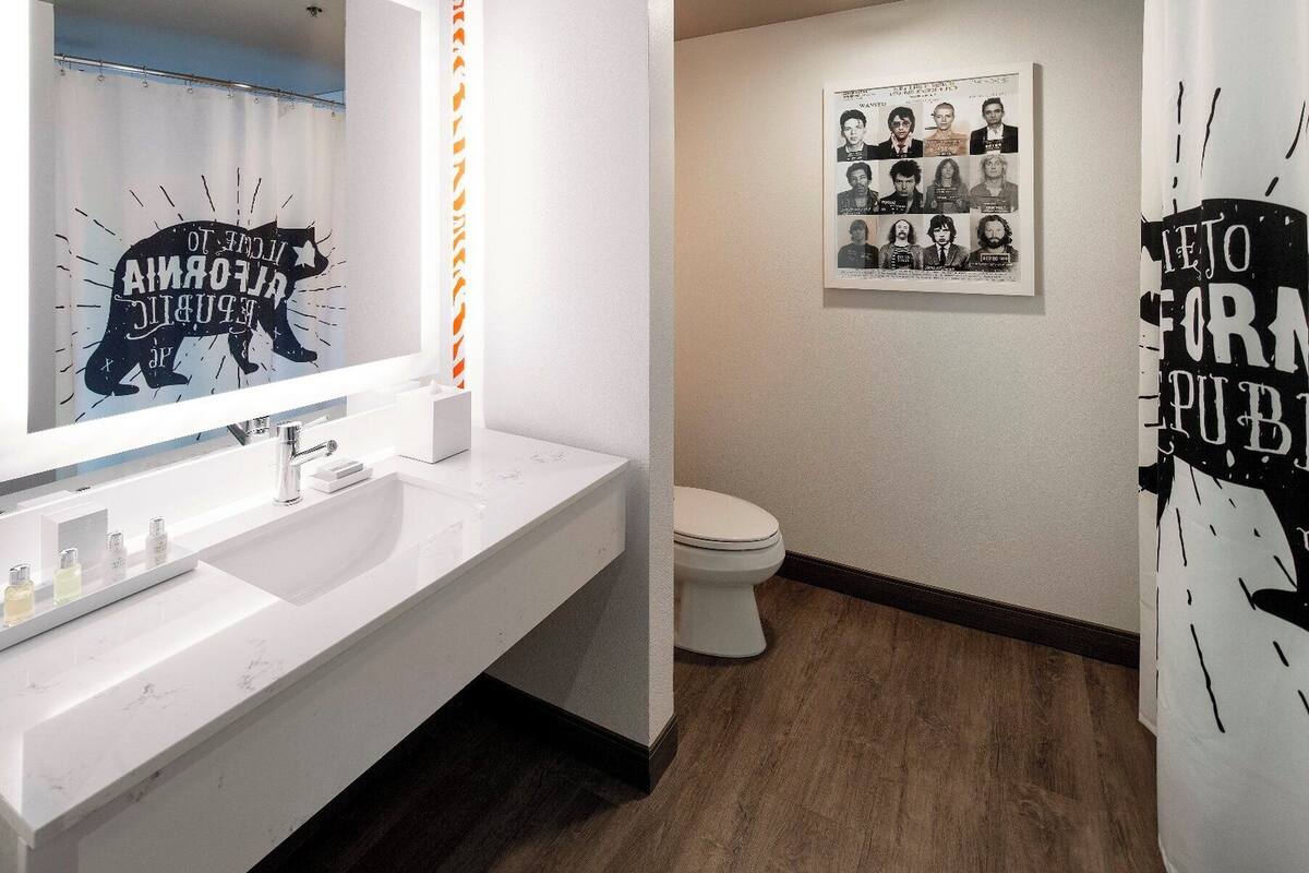 Ein modernes Badezimmer mit einem weißen Waschbecken, Spiegel und Toilette. An der Wand befinden sich Poster und ein Duschvorhang mit Schrift.