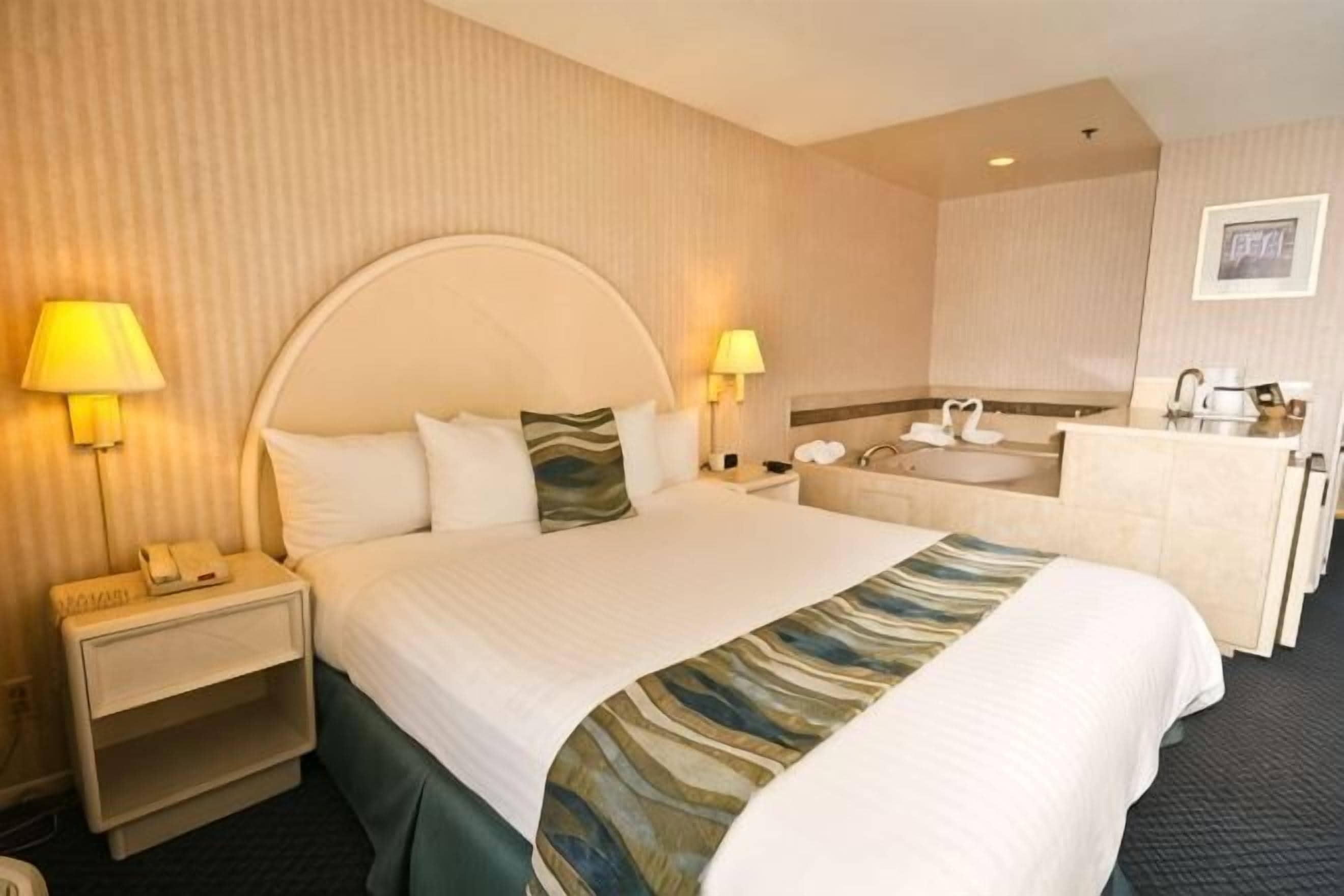 Ein Hotelzimmer mit einem großen Bett, zwei Nachttischlampen, einem Nachttisch, einem Schreibtisch, einem Badezimmer mit Waschbecken und Spiegel sowie einem gerahmten Bild an der Wand.