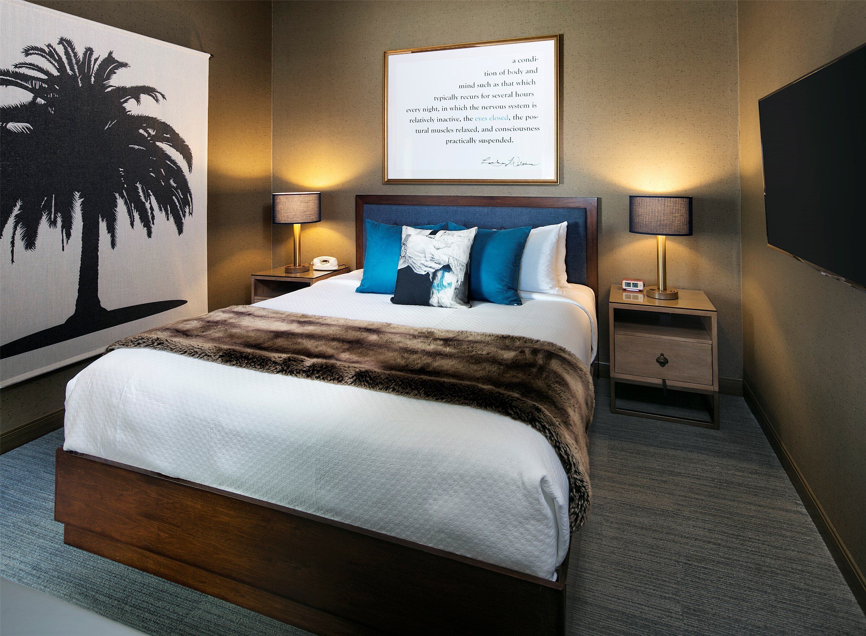 A neatly arranged hotel room with a large bed, two bedside tables with lamps, a framed quote on the wall, and a large painting of a palm tree.