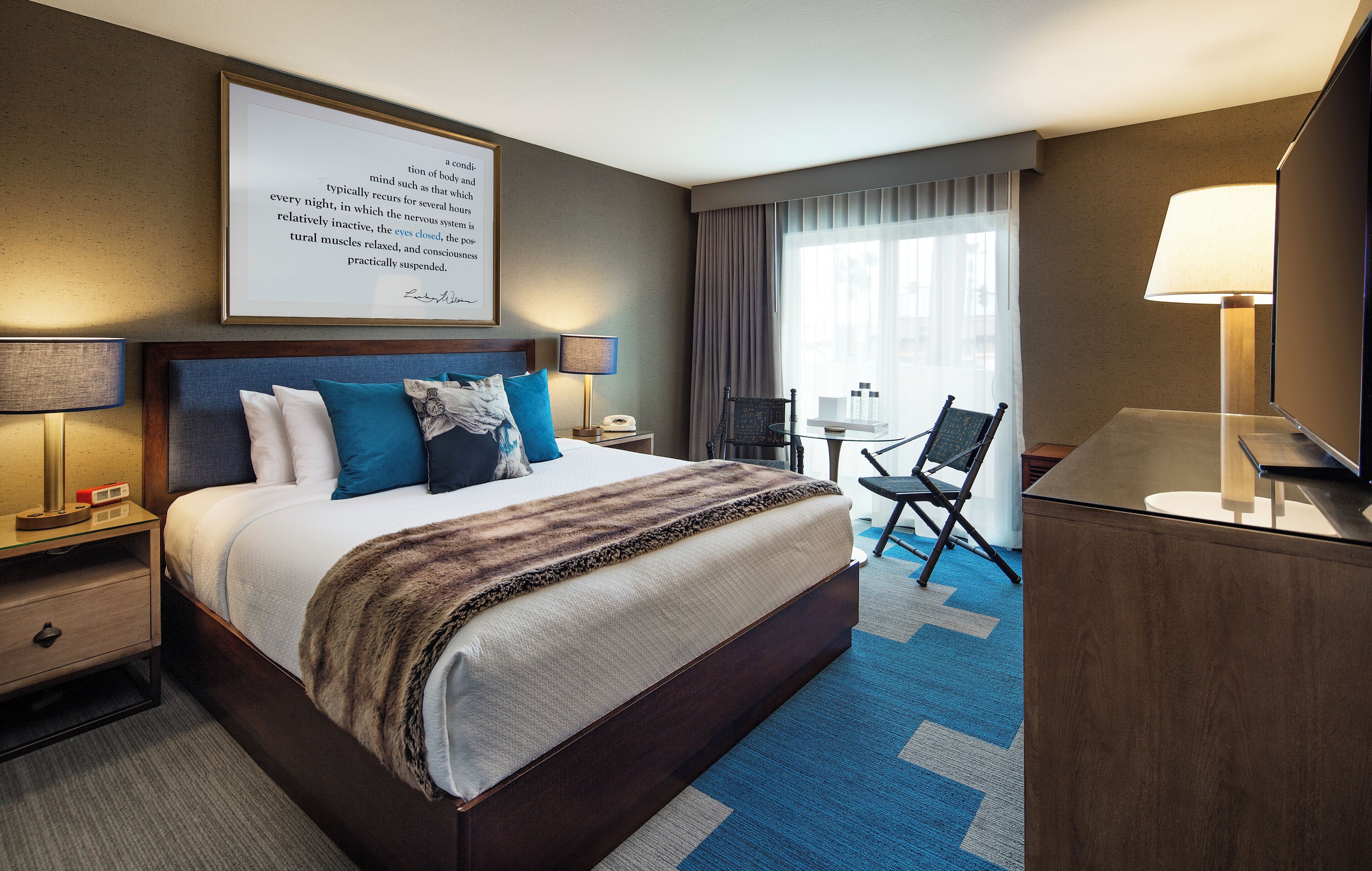 A hotel room with a large bed, bedside tables, a desk, a chair, and a window with curtains.