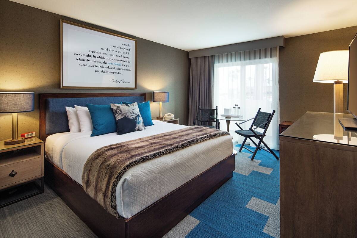 A hotel room with a large bed, bedside tables, a desk, a chair, and a window with curtains.