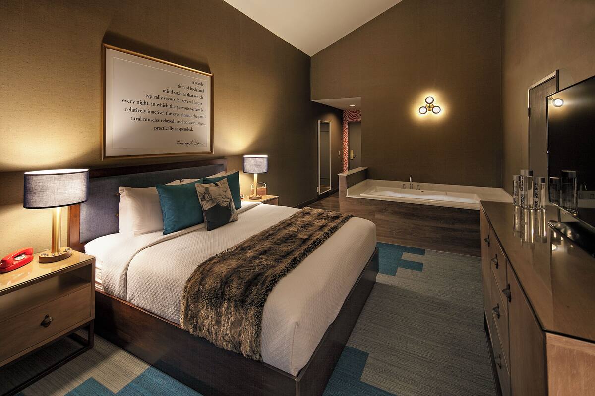 A modern hotel room with a large bed, a bedside table with a lamp, a nightstand, a dresser, a large mirror, and a framed quote on the wall.