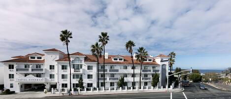 A multi-story hotel with a white facade and red-tiled roof, featuring balconies and palm trees.