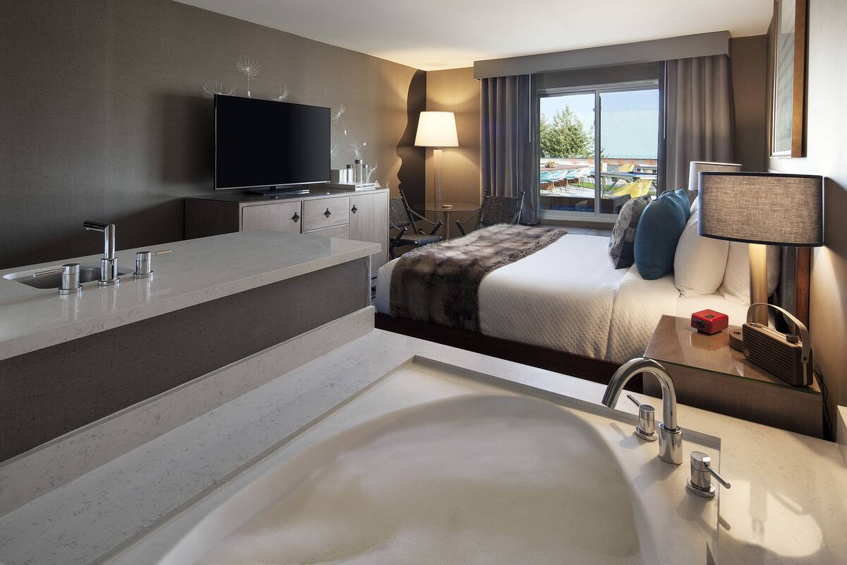 A modern hotel room with a large bed, a flat-screen TV, a large window with a view, a bathroom with a bathtub, and a desk with a lamp.