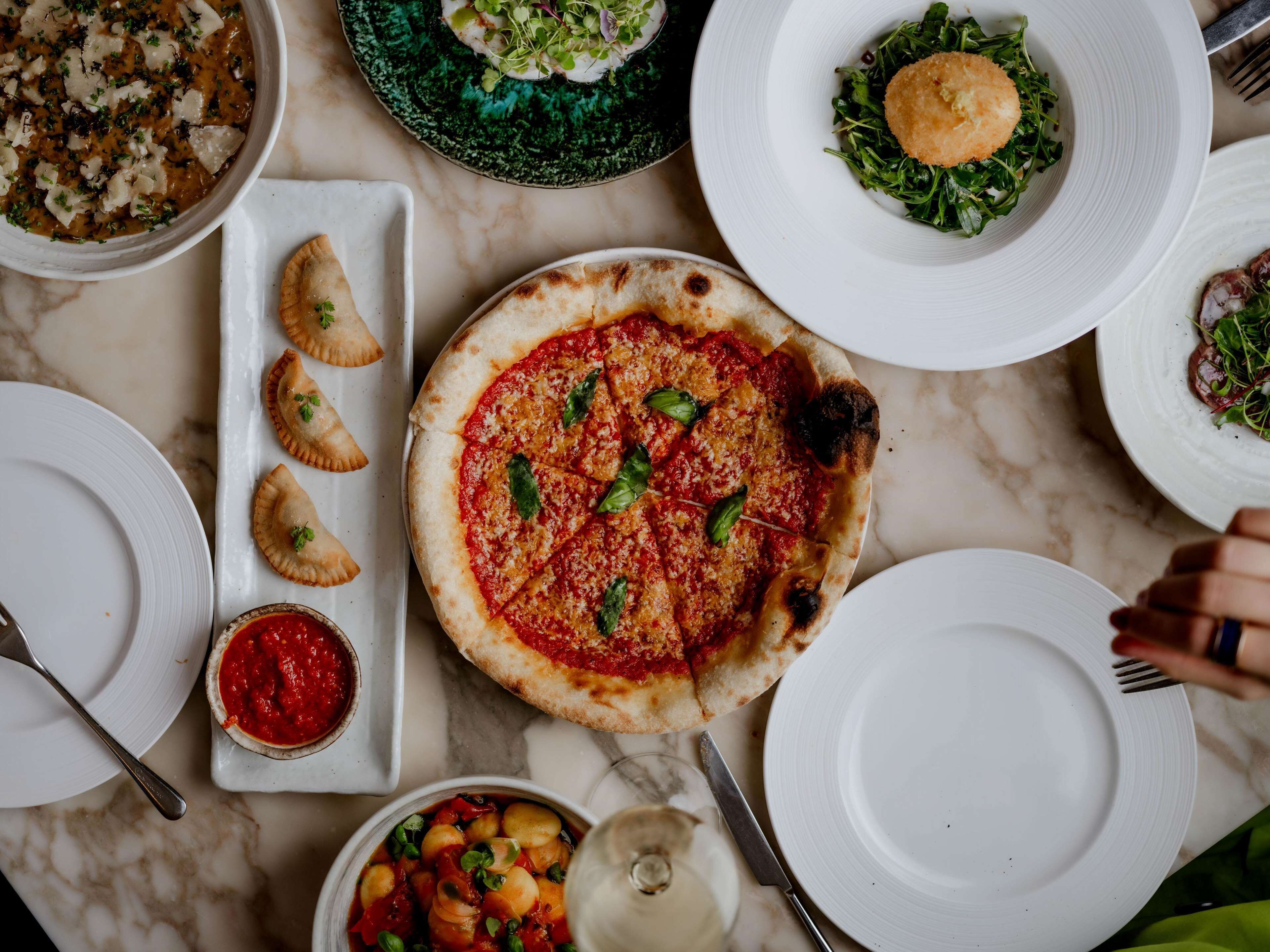 A variety of dishes including pizza, ravioli, and a bowl of soup, arranged on a marble table.