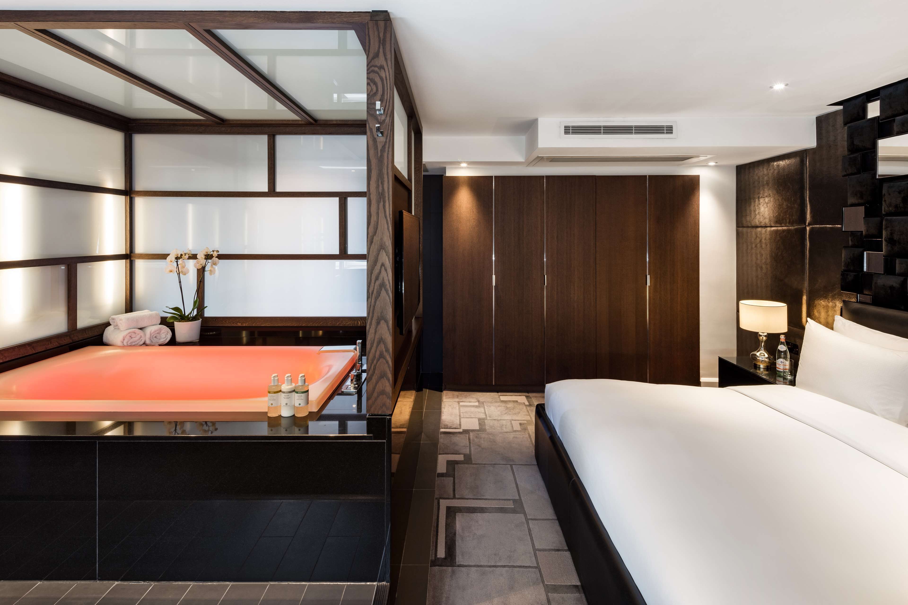 A modern hotel room with a large bed, a glass-enclosed shower area, a wooden wardrobe, and a nightstand with a lamp.