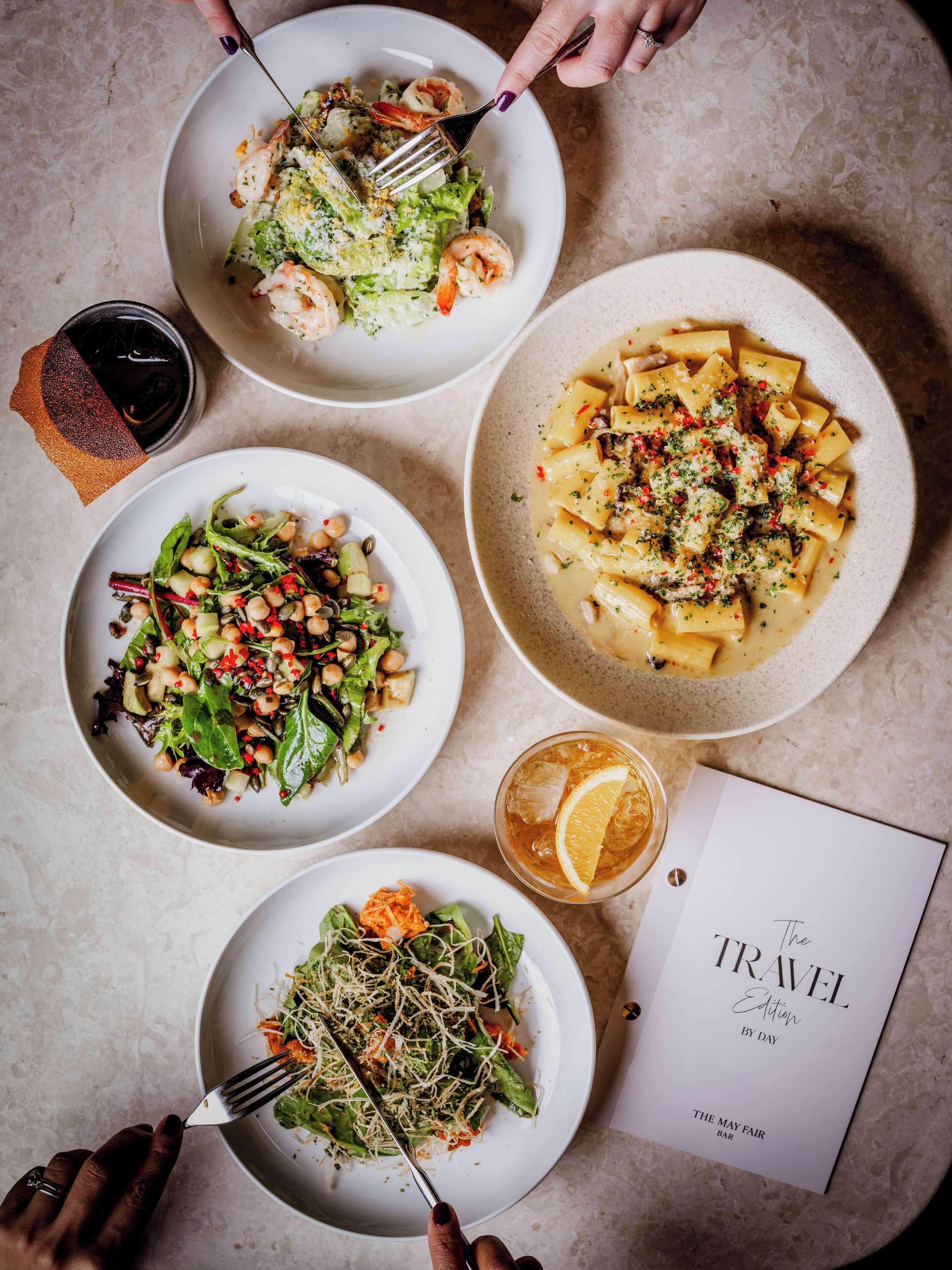 A table with four plates of food, a glass of iced tea, and a menu titled "The Travel Edition by Day".
