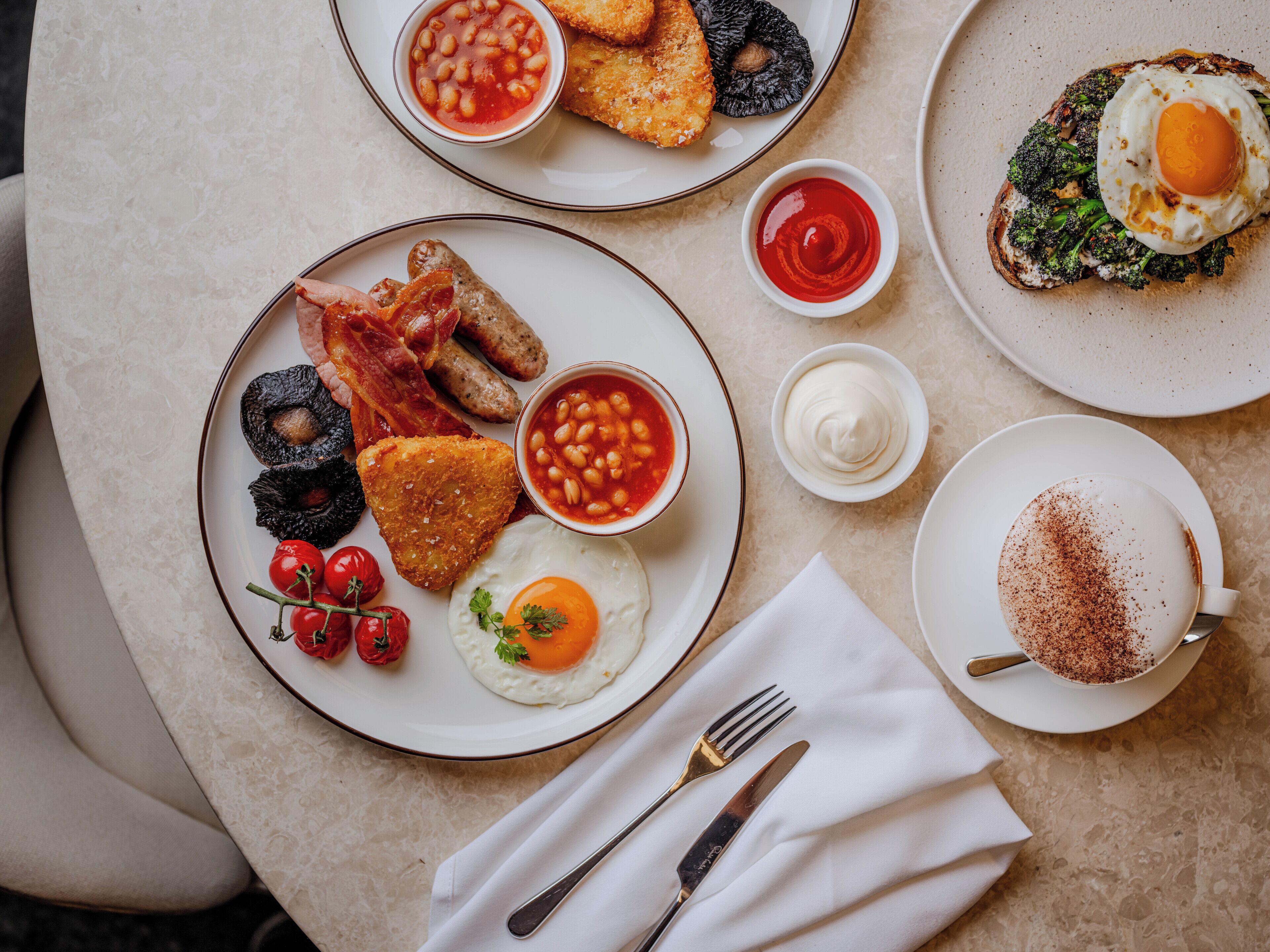 Una colazione con uova, pancetta, salsicce, toast, fagioli e una tazza di caffè su un tavolo.