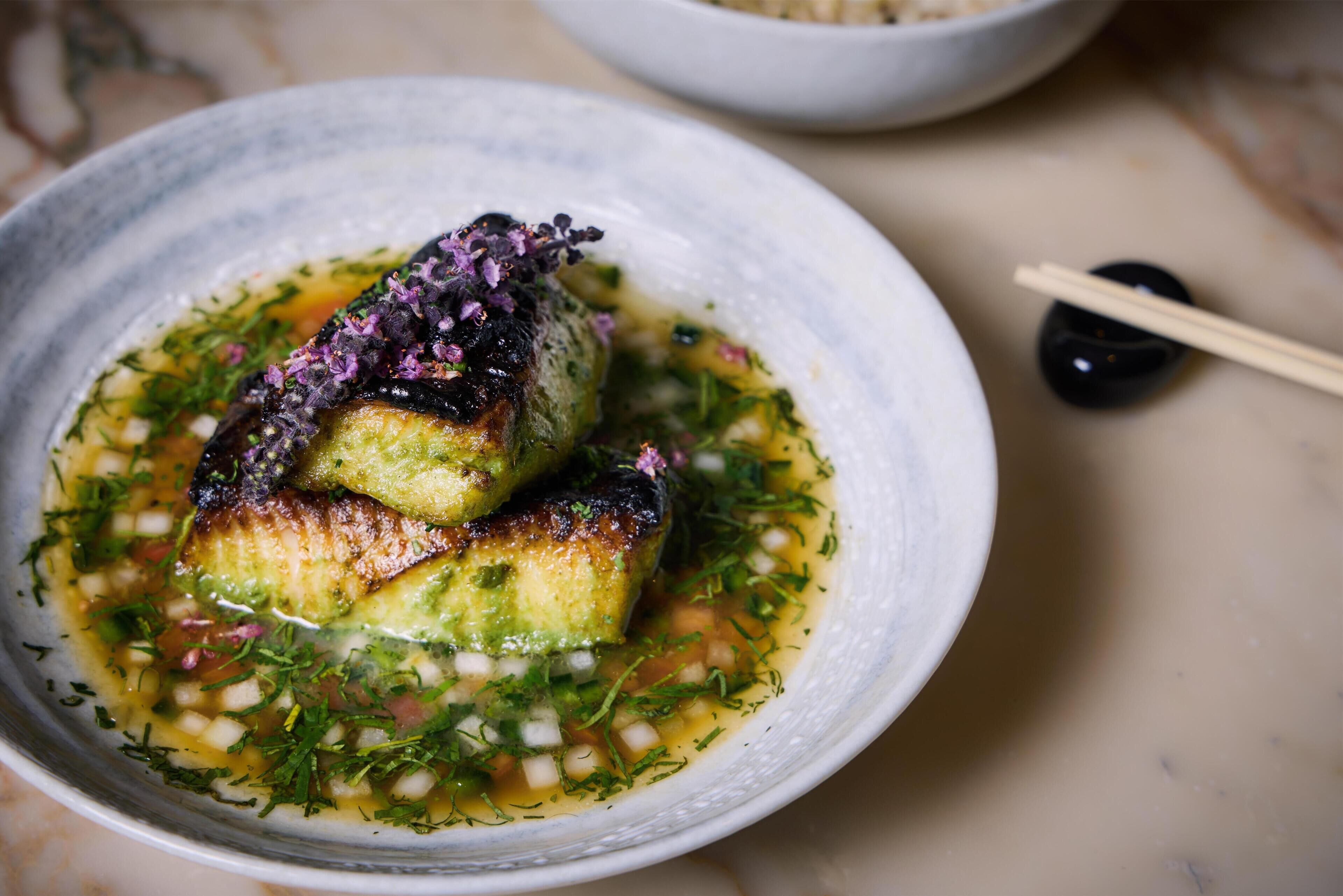A bowl of food with a fish and greens, a pair of chopsticks, and a small black object.