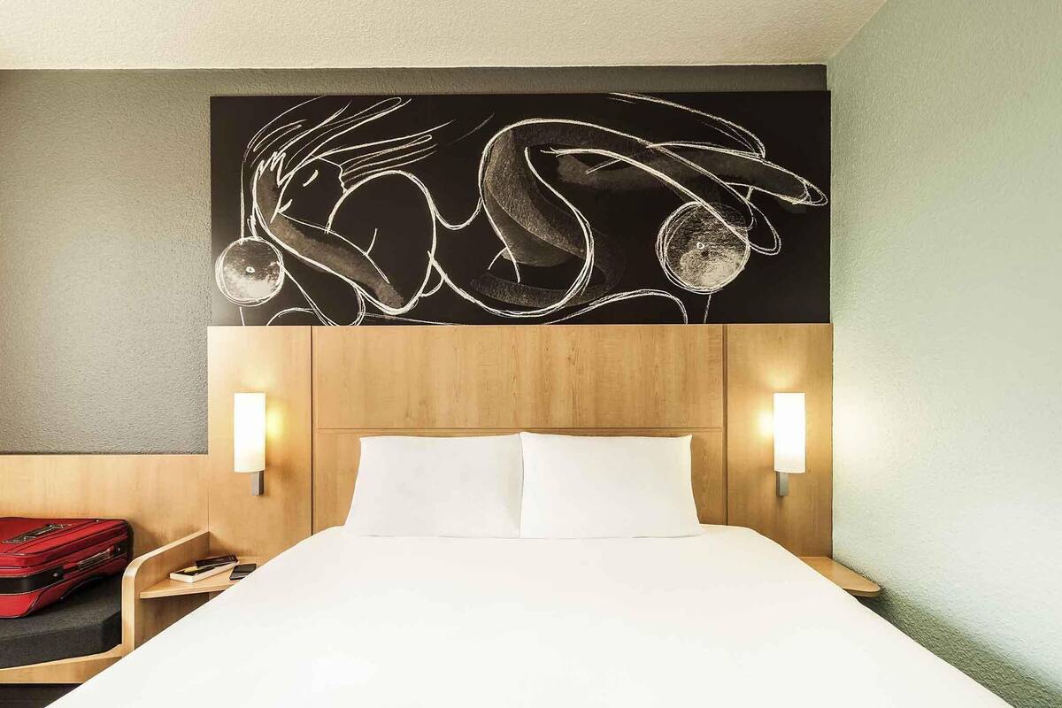 A hotel room with a bed, a red suitcase, a nightstand, and a wall art piece.