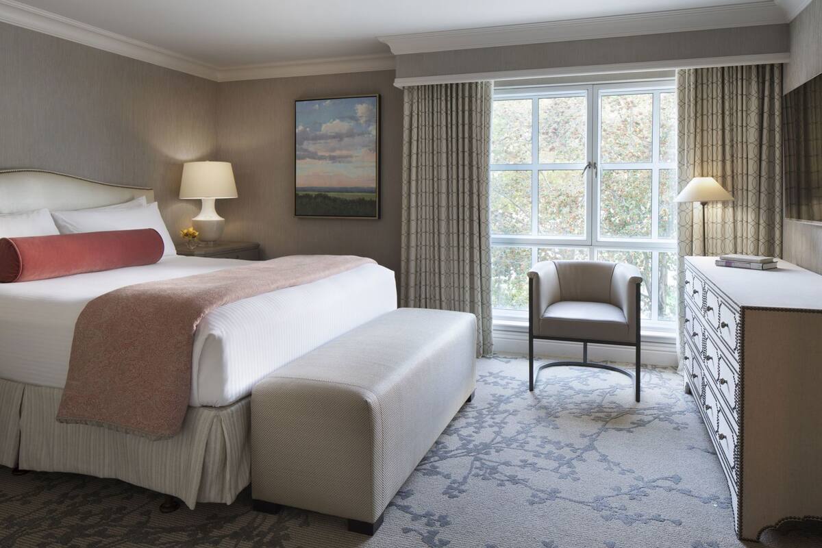 Una habitación de hotel con una cama grande, mesitas de noche, una silla, un aparador y una ventana con cortinas.