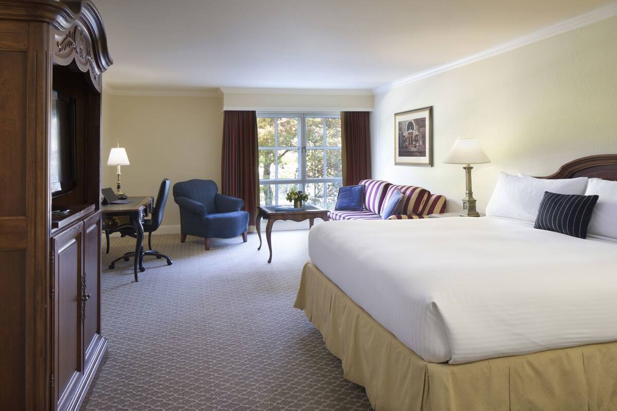 Habitación de hotel con una cama grande, un escritorio con silla, televisión y ventana con cortinas.