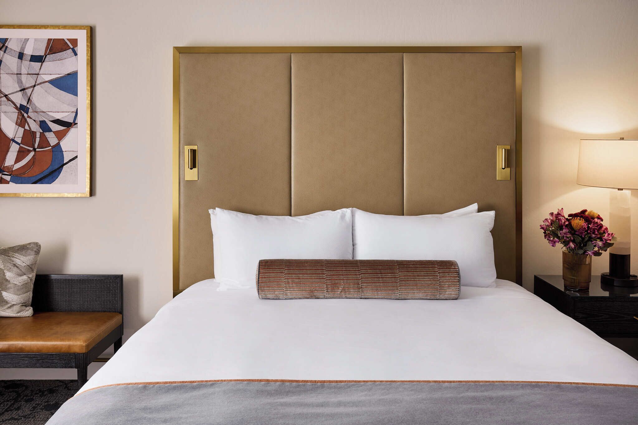 A hotel room with a large bed, a headboard, a bedside table with a lamp, a chair, and a painting on the wall.