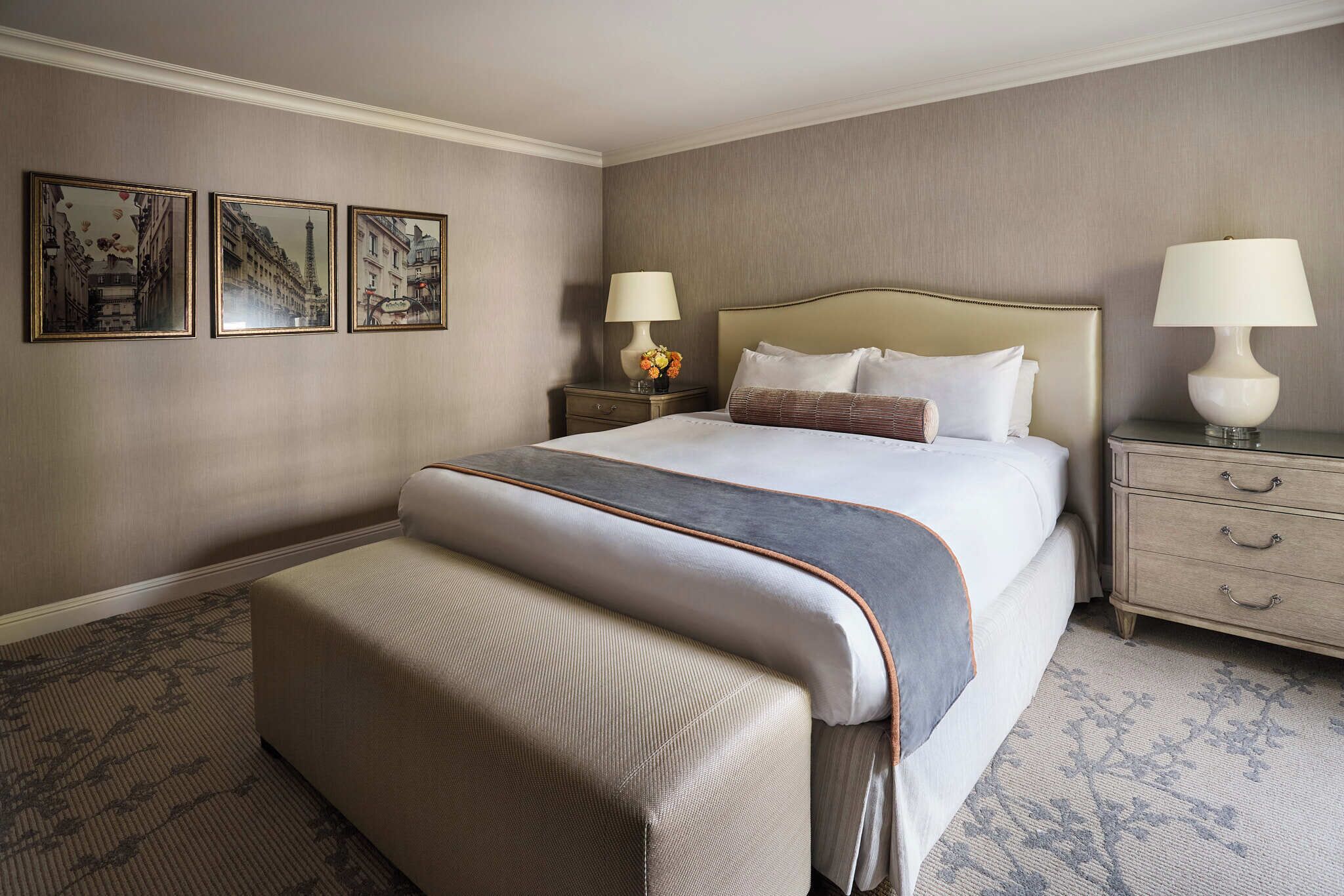 Une chambre d’hôtel avec un grand lit, deux tables de chevet avec des lampes, un banc au pied du lit et trois tableaux encadrés au mur.