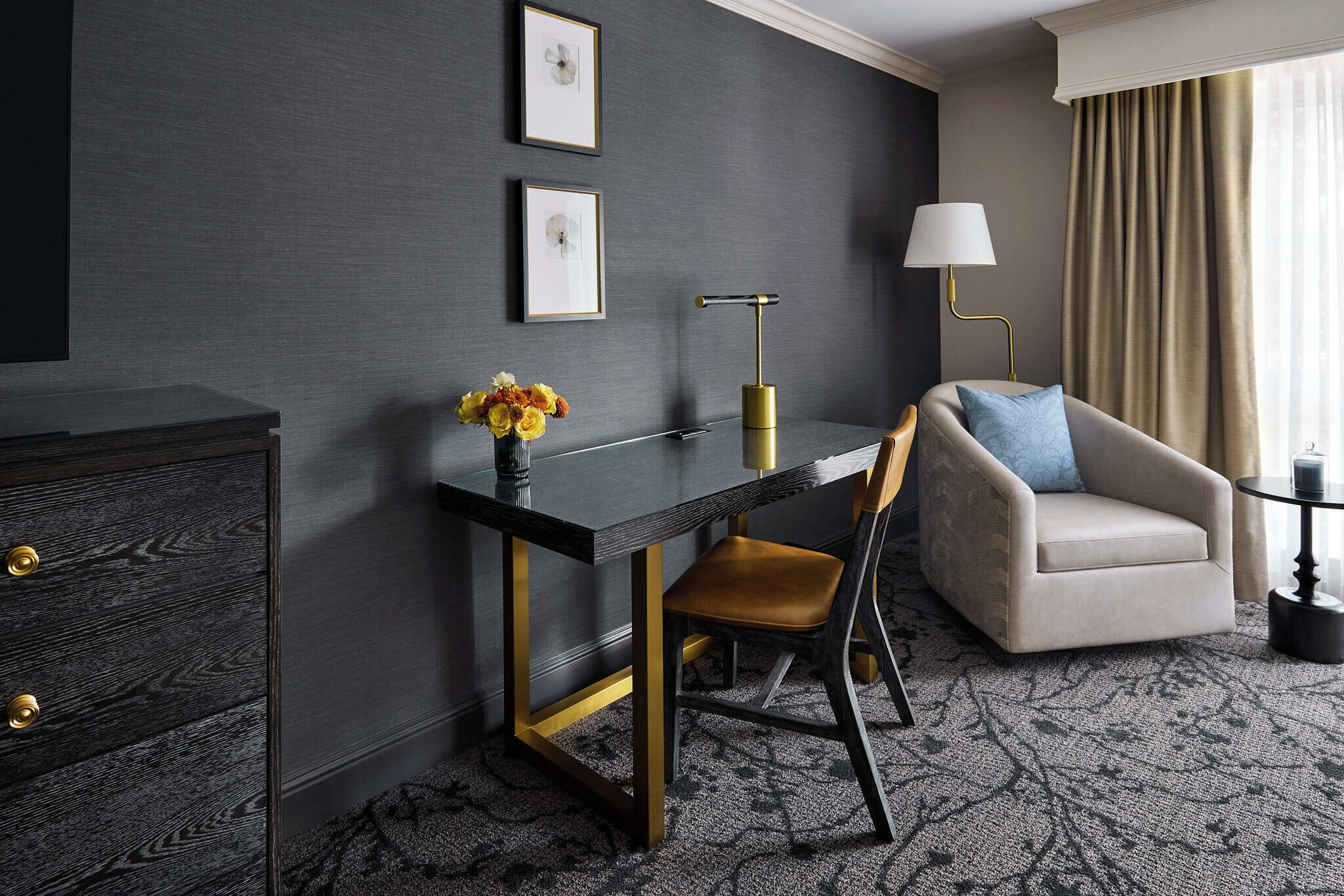A modern hotel room with a dark grey wall, a black desk with gold accents, a beige armchair, a small side table with a lamp, and a vase with flowers.