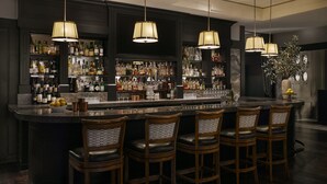 Un espace bar aux couleurs sombres, avec de nombreuses bouteilles d’alcool et quatre tabourets de bar.