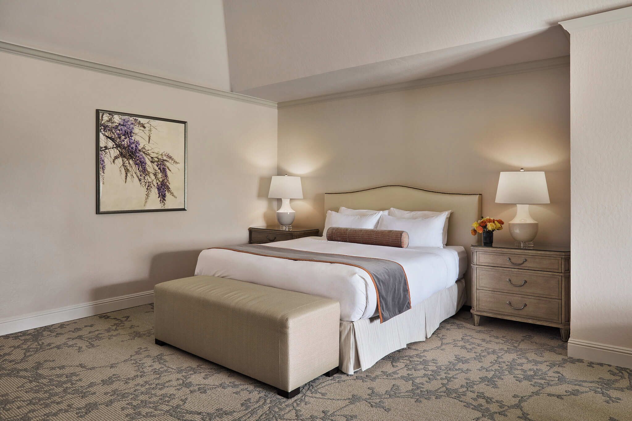 A neatly arranged bedroom with a large bed, two bedside tables with lamps, a bench, and a floral artwork on the wall.
